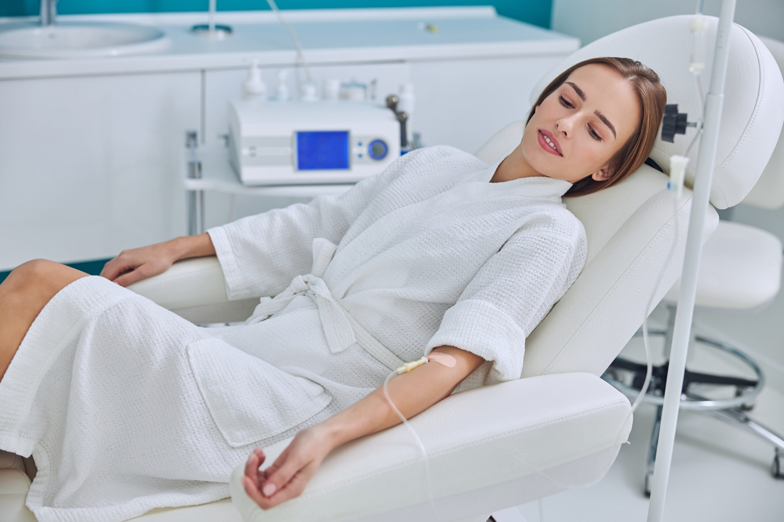 Waist up side view portrait of young pretty lady in white bathrobe lying on medicine armchair while receiving vitamin medical procedure in wellness center